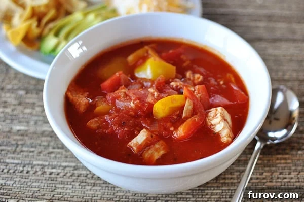 A beautifully presented white bowl full of red chicken chili, with a selection of enticing toppings like cheese, onions, and avocado on a white plate in the background, ready for customization.