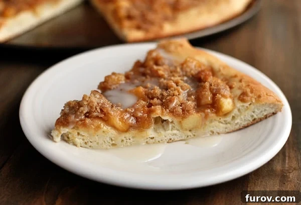 Golden Apple Streusel Dessert Pizza 4 A tempting slice of apple cinnamon dessert pizza, beautifully garnished with a thick, white vanilla glaze, on a white ceramic plate.