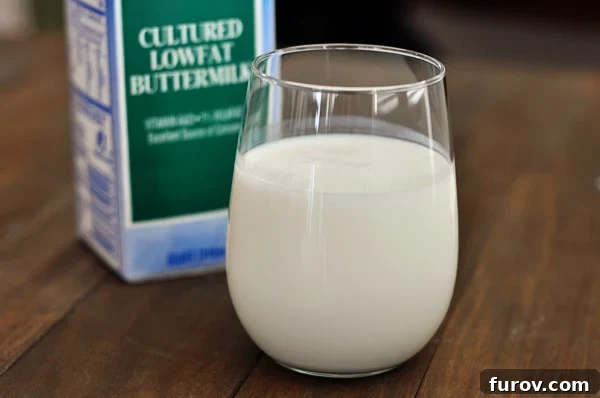 A clear glass of fresh buttermilk with a carton of buttermilk positioned behind it, illustrating the liquid's texture and color.