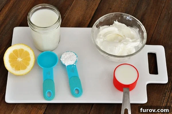 Measuring spoons, a fresh lemon, a glass of milk, and a bowl of sour cream arranged on a white cutting board, illustrating ingredients for various buttermilk substitutes.