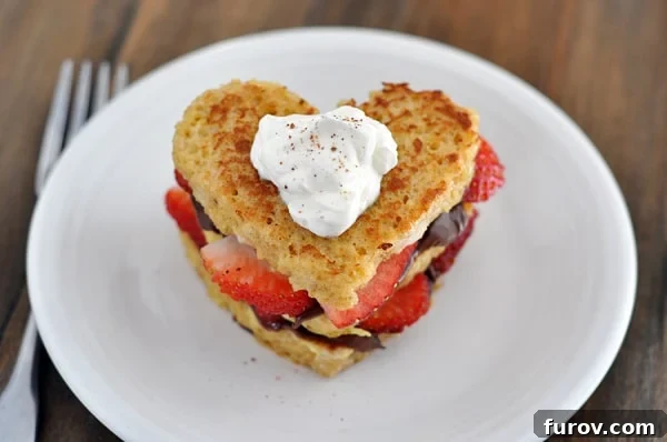 Top view of a heart shaped chocolate and strawberry stuffed french toast on a white plate, garnished with whipped cream and a sprinkle of nutmeg.