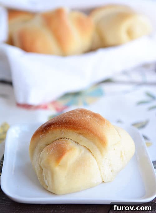 Fluffy Buttermilk Rolls: Prep Now, Enjoy Later 3 A golden brown buttermilk roll resting on a white plate, with a basket of more freshly baked rolls blurred in the background.