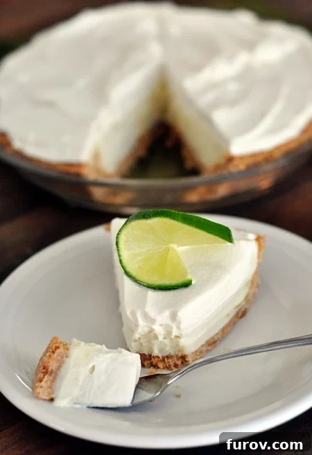 A generous slice of sour cream lime tart with its golden graham cracker crust, presented on a white plate with the rest of the pie blurred in the background.