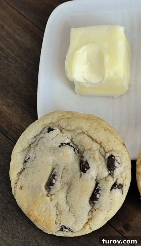 A close-up, top-down view of a perfectly formed and baked chocolate chip cookie, boasting ideal texture and even browning, accompanied by a stick of butter at the perfect, cool room temperature.