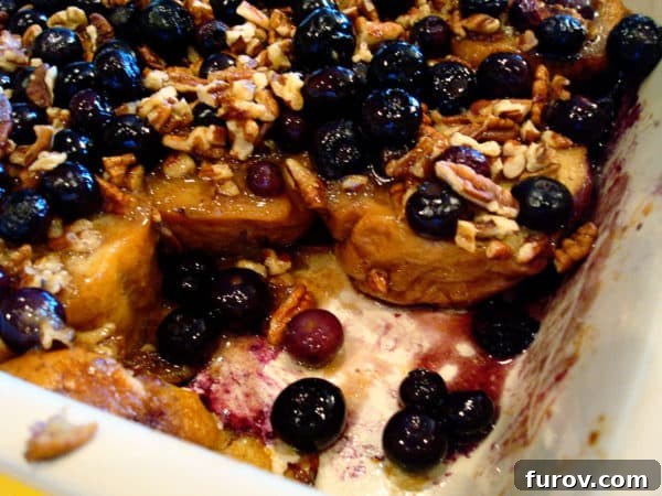 A serving of baked French toast with blueberries and pecans removed from a white dish, showcasing the golden-brown crust and juicy interior.