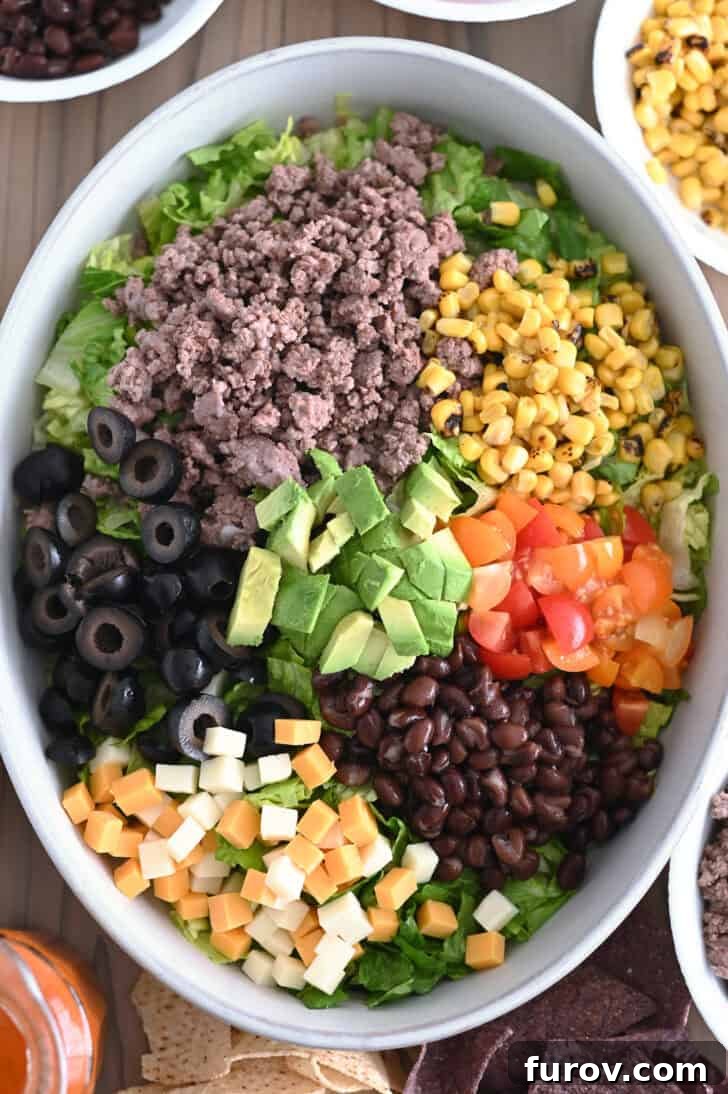 Zesty Chopped Taco Salad with Homemade Catalina 4 Top down view of bowl with lettuce, black beans, chopped tomatoes, roasted corn, cooked ground beef, sliced olives, cubed cheese and avocados, partially assembled.