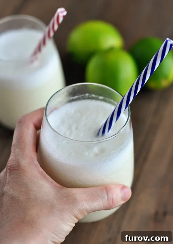 Velvet Brazilian Lemonade 2 A glass of frothy lemonade with a blue and white striped straw in it and limes in the background, exuding a fresh and inviting appeal.