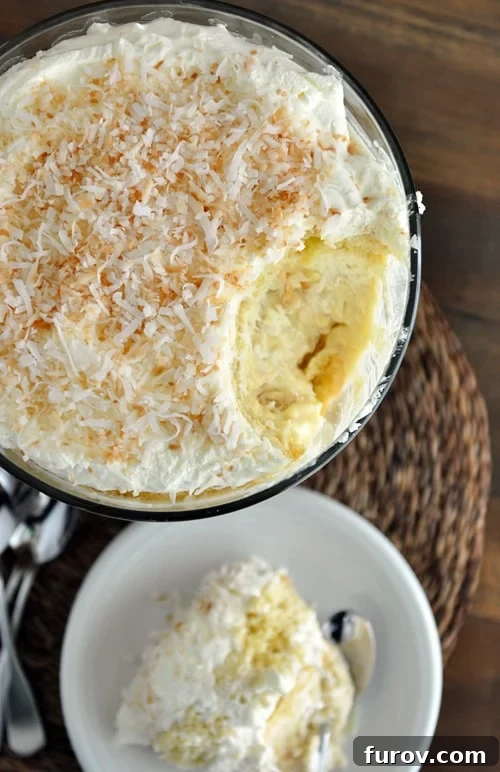 Decadent Coconut Tres Leches Trifle 3 Top-down view of a Coconut Tres Leches Cake Trifle in a glass bowl, with a scoop removed to reveal the exquisite layers beneath, garnished with toasted coconut.