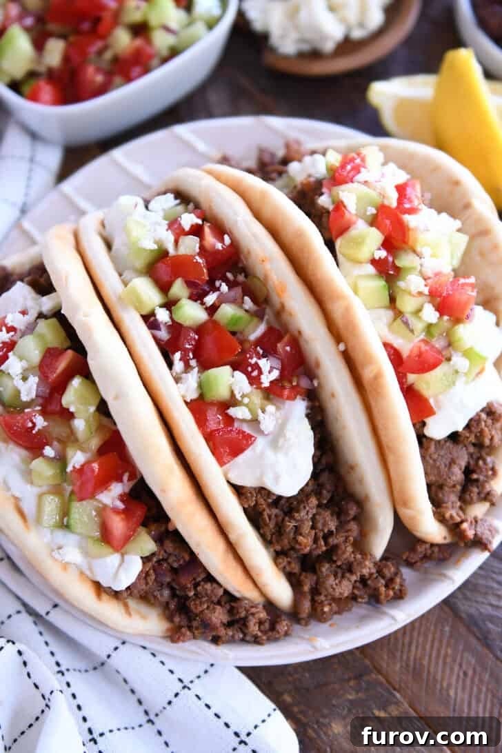 Olympian Taco Feast 2 Top down view of three loaded Greek flatbread tacos, generously topped with creamy tzatziki sauce, crisp diced cucumbers, and juicy chopped tomatoes, on a rustic wooden surface.