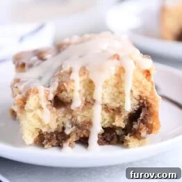 Square of cinnamon roll cake on white plate with glaze on top and sides.