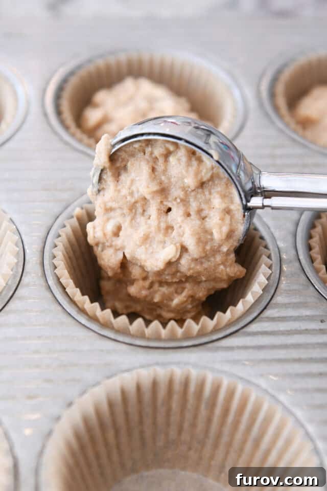 A hand using a cookie scoop to portion the applesauce oat muffin batter into a lined muffin tin, ensuring even sizes.