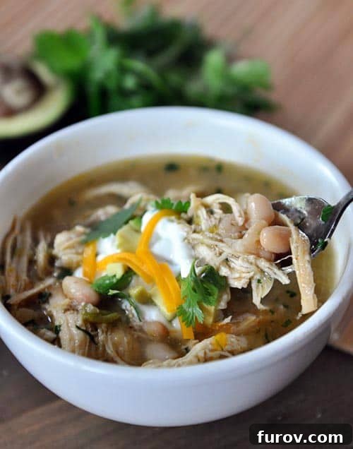 A steaming white bowl of white bean chicken chili with a spoon taking a bite out, showcasing the creamy texture and tender chicken.