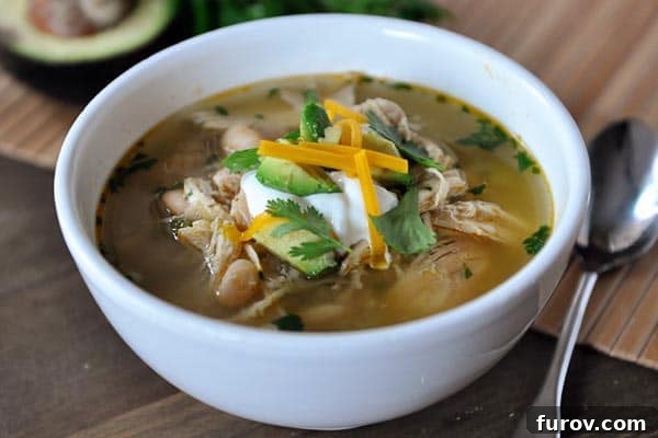 Close-up of a white bowl filled with slow cooker white bean chicken chili, generously topped with creamy sour cream, vibrant diced avocado, and shredded cheese.