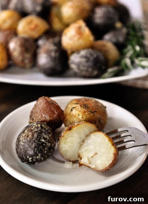 A white plate featuring four cooked baby potatoes, one halved to show its creamy interior, with a larger platter of potatoes in the background.