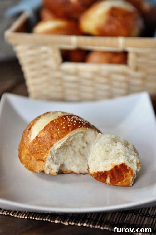Pillowy Soft Pretzel Buns 4 A golden brown pretzel roll with a chunk split off, resting on a white plate, showcasing its soft interior.