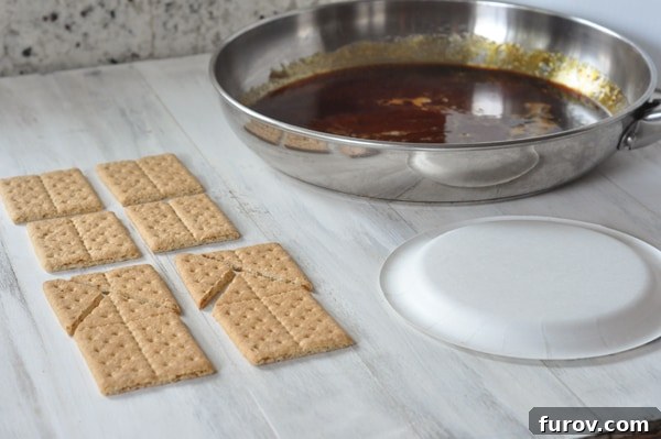 A workstation set up with graham cracker pieces, a white plate, and a pan of melted, shimmering sugar, ready for house assembly