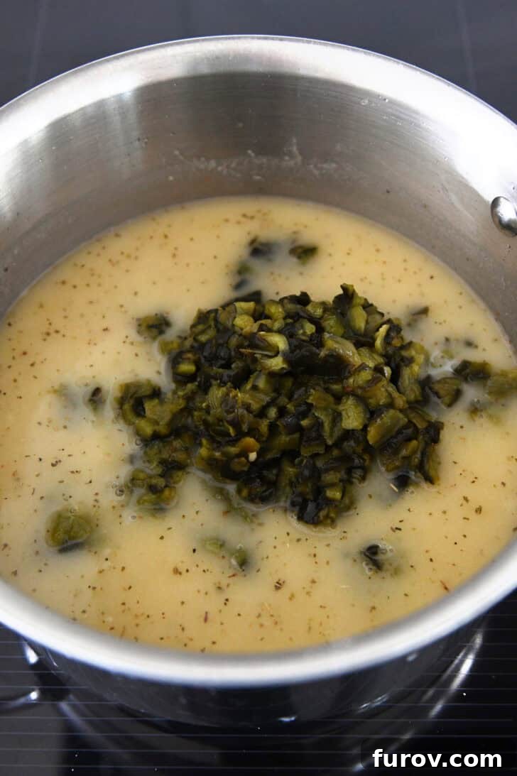 A metal pot filled with a simmering broth mixture, with roasted, diced green peppers piled on top, ready to be incorporated.