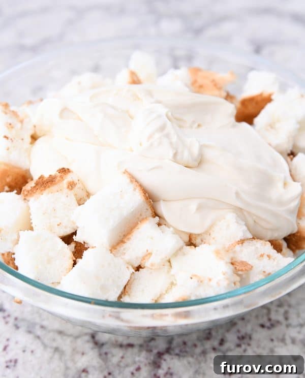 A large mixing bowl containing cubed angel food cake pieces, being generously coated with a smooth, creamy mixture.