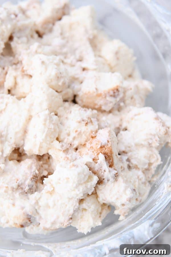 A clear glass bowl showcasing numerous angel food cake pieces, perfectly and thickly coated with the cream cheese mixture, ready for the next layer.