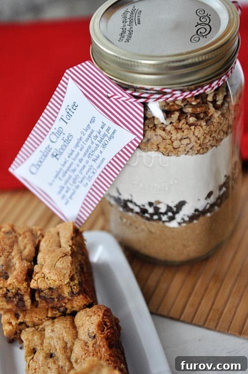 Blondie Indulgence Jar 2 A beautifully layered mason jar containing dry ingredients for chocolate chip toffee blondies, with baked blondies arranged artfully on the side, highlighting its appeal as a gift.