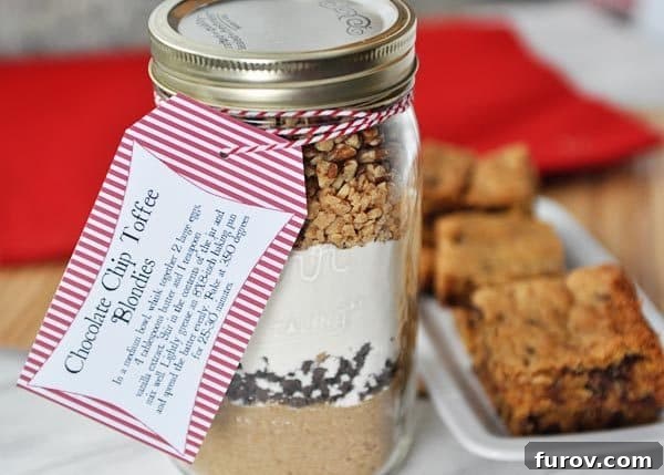 Blondie Indulgence Jar 4 A clear glass mason jar filled with distinct layers of dry ingredients for chocolate chip toffee blondies, positioned next to a platter of freshly baked blondies. An excellent homemade gift idea.