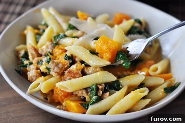 A close-up of a white bowl filled with creamy penne pasta, tender butternut squash, flavorful sausage, and vibrant spinach, garnished with fresh herbs.