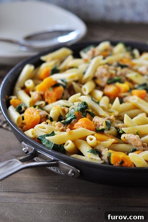 Skillet full of perfectly cooked penne pasta, tender cubes of butternut squash, fresh spinach, and savory cooked sausage, ready for the final cheese melt.