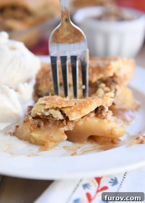 Golden Toffee Crunch Caramel Apple Pie 4 A fork taking a bite of Toffee Crumble Caramel Apple Pie from a white plate, accompanied by a scoop of creamy vanilla ice cream.