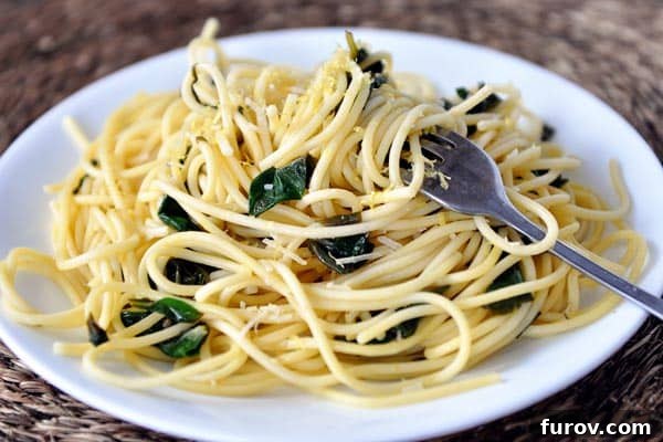 White plate with cooked spaghetti with spinach sprinkled throughout, garnished with lemon wedges.
