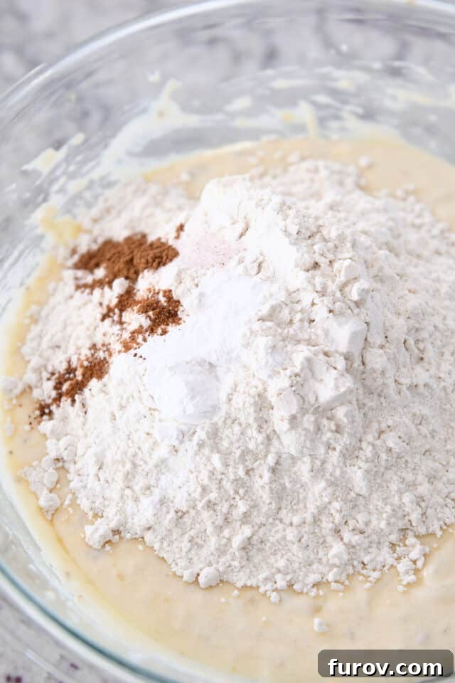 Flour, cinnamon, salt and baking powder being folded into the banana bread batter in a glass bowl.
