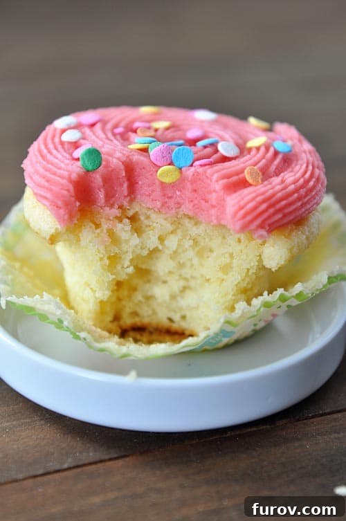 A vibrant yellow cupcake with fluffy pink frosting and colorful sprinkles, with a bite taken out, resting on a small white dish.