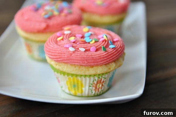 A pristine white platter adorned with several yellow cupcakes, each topped with a swirl of pink frosting and colorful sprinkles, ready for serving.