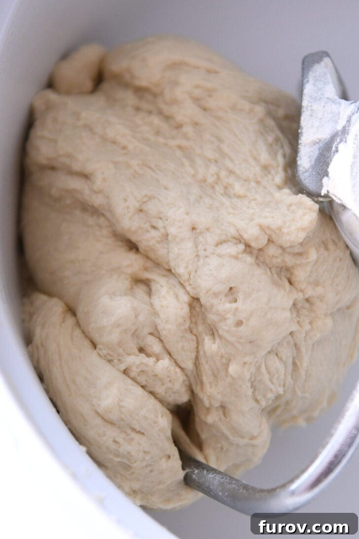 Top down view of a stand mixer bowl with a dough hook, showing a large, perfectly formed ball of bread dough, smooth and ready for the next stage.