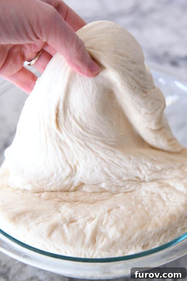 A hand gently pulling soft, elastic bread dough out of a glass mixing bowl, demonstrating its ideal consistency.