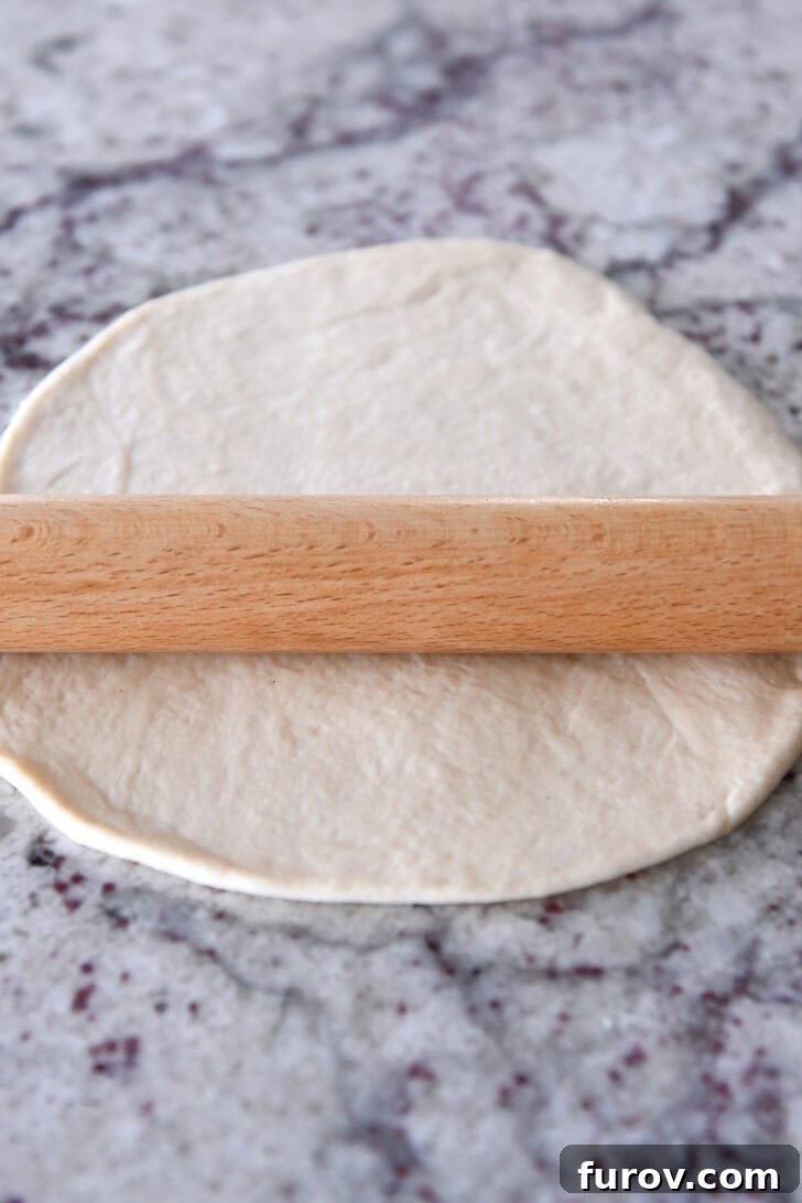 A wooden rolling pin smoothly rolling out a piece of bread dough into a thin, even circle on a granite countertop, showcasing the ease of shaping.