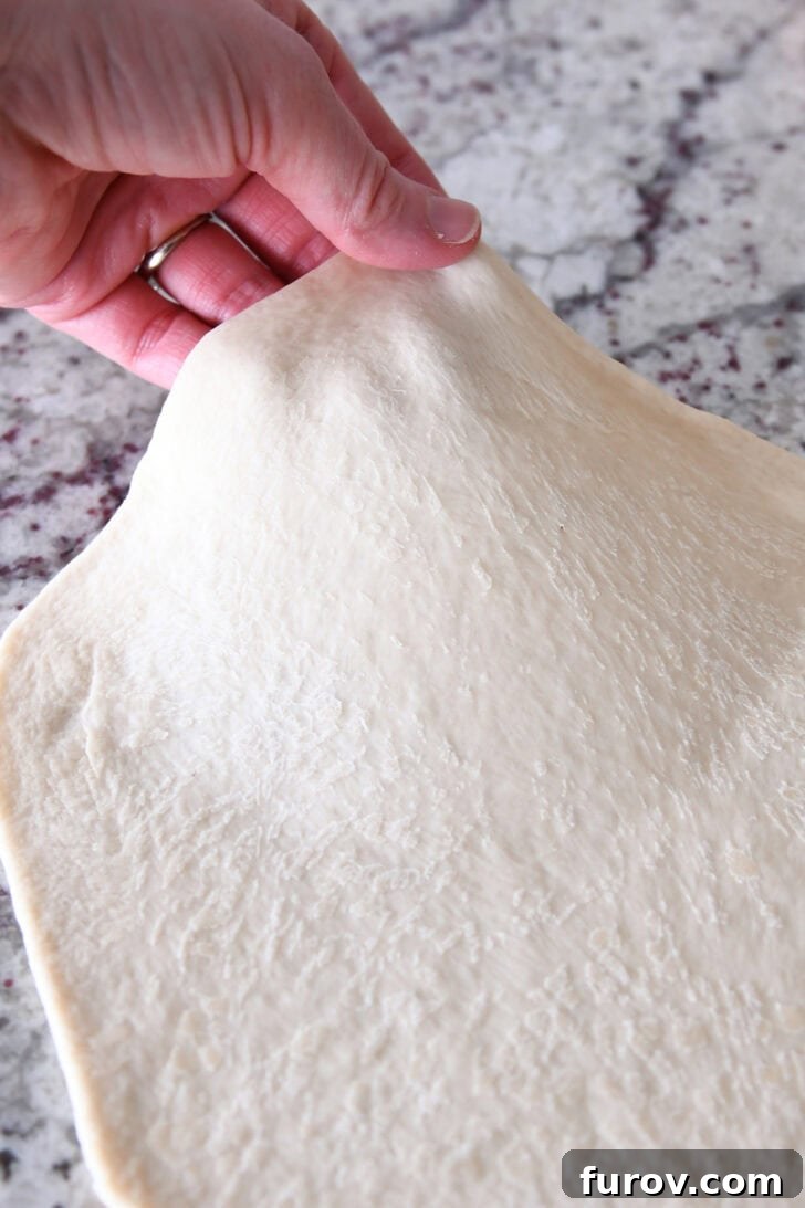 A hand gently lifting the edge of a rolled-out piece of bread dough from the counter, showing its flexibility before cooking.
