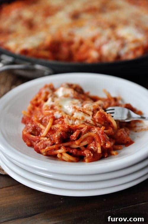 A stack of pristine white plates, with the top plate showcasing a generous, steaming serving of cheesy skillet baked spaghetti, garnished with fresh parsley.