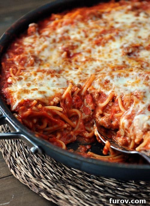 A robust cast iron skillet filled to the brim with bubbly, golden cheese-topped baked spaghetti, fresh from the oven and ready to be served.