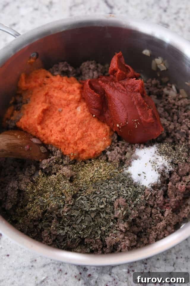 Pot with ground beef, basil, oregano, salt, tomato paste, and blended red pepper and onion, sizzling on the stove.