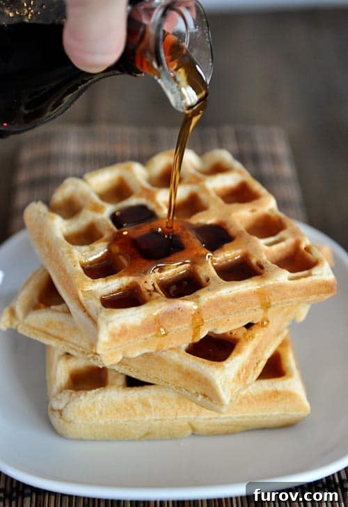 A hand pouring syrup over a stack of thick, golden-brown overnight raised waffles, highlighting their fluffy texture.