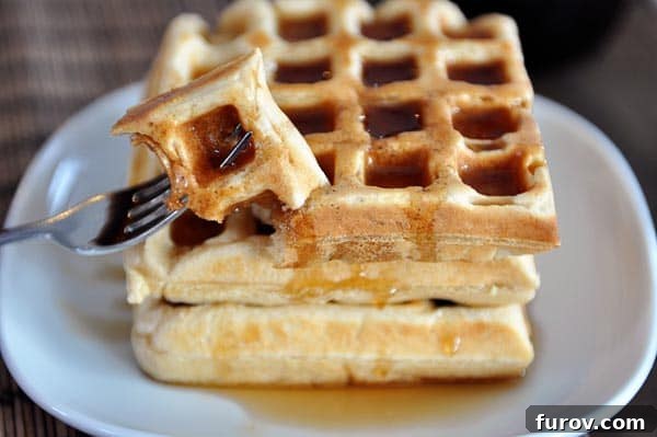 A stack of golden-brown, fluffy overnight waffles, generously covered in maple syrup, served on a pristine white plate.