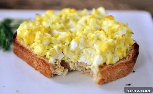 An open-faced egg salad sandwich with a bite taken out, sitting on a white plate, showing the warm, slightly toasted top.