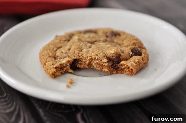 A white plate featuring a delicious whole wheat coconut oil chocolate chip oatmeal cookie with a bite taken out, revealing its soft, chewy center and melted chocolate.