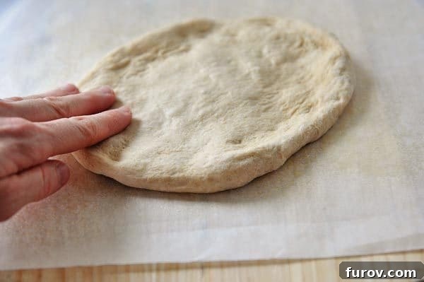 Fast and Flawless Pizza Dough 10 Uncooked pizza dough being flattened on parchment paper, ready to be topped.