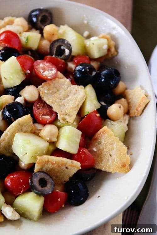 Top view of a white bowl of Greek pita salad filled with toasted pitas, crisp cucumbers, briny olives, ripe tomatoes, and wholesome chickpeas.