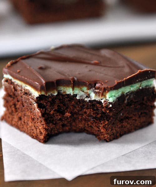 A chocolate frosted mint brownie with a bite taken out, sitting on a square piece of parchment paper. The rich layers of chocolate, vibrant mint, and glossy ganache are visible.