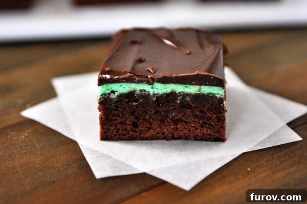 A perfectly cut layered mint brownie showing off its distinct layers of chocolate, green mint frosting, and dark chocolate ganache, resting on two squares of parchment paper.