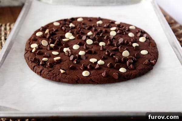 A giant white and chocolate chip topped chocolate cookie on a sheet of parchment paper, ready to be enjoyed.