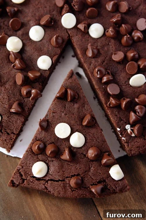 A pie-shaped piece cut out of a giant chocolate chip studded chocolate cookie, ready to be served.