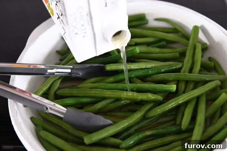 Pouring chicken broth into a hot skillet with fresh green beans and tongs, illustrating a key step in making easy skillet green beans.